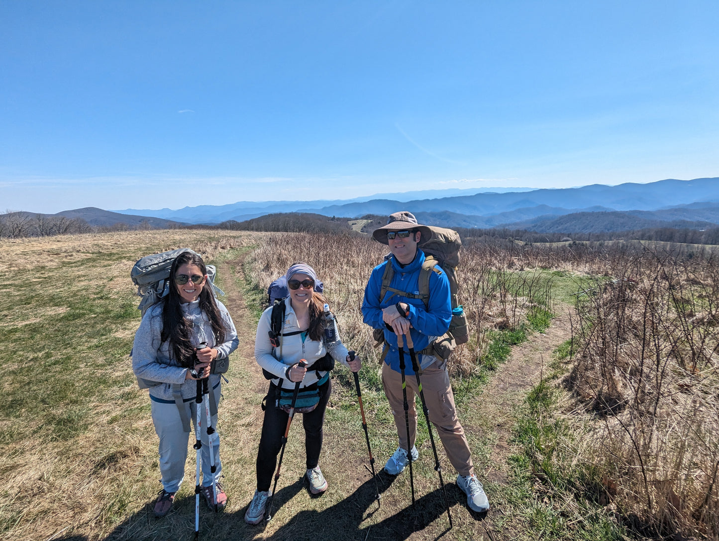 Two women and a man with backpacks and trekking poles standing on a hiking trail with mountains in the distance