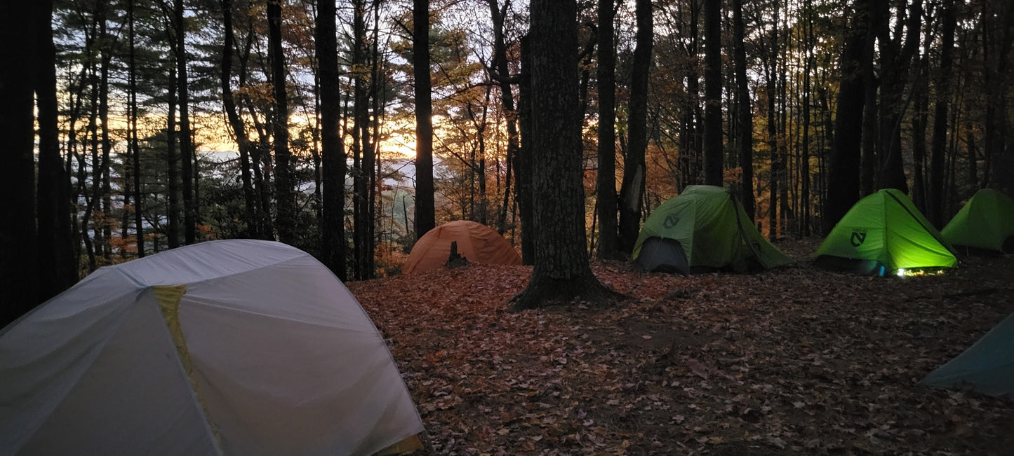 A group of backpacking tents set up in the woods as the sun sets in the distance