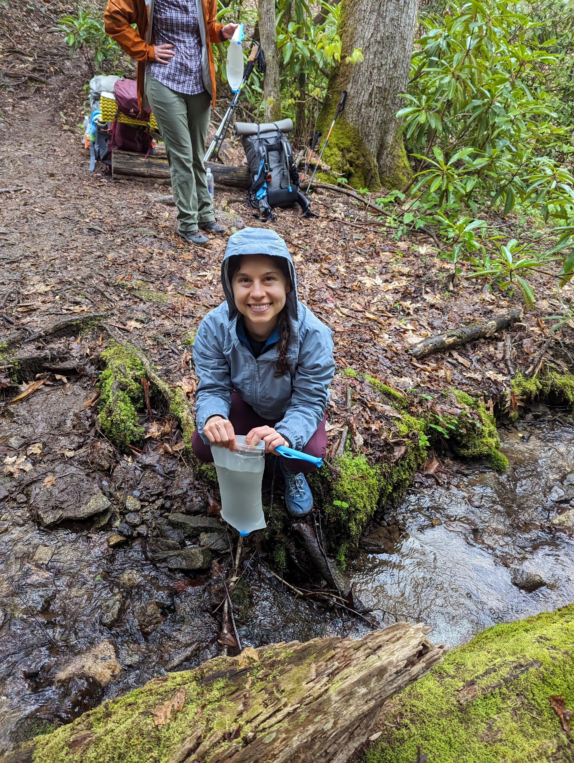 A young woman fills a water filter bag at a stream next to a hiking trail