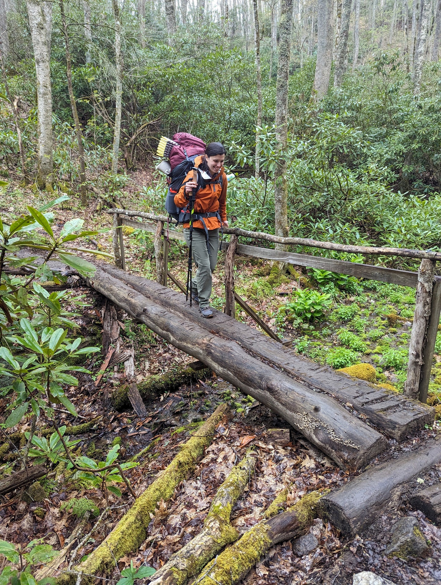 A woman wearing a backpack and rain jacket walks across a wooden log bridge in the forest