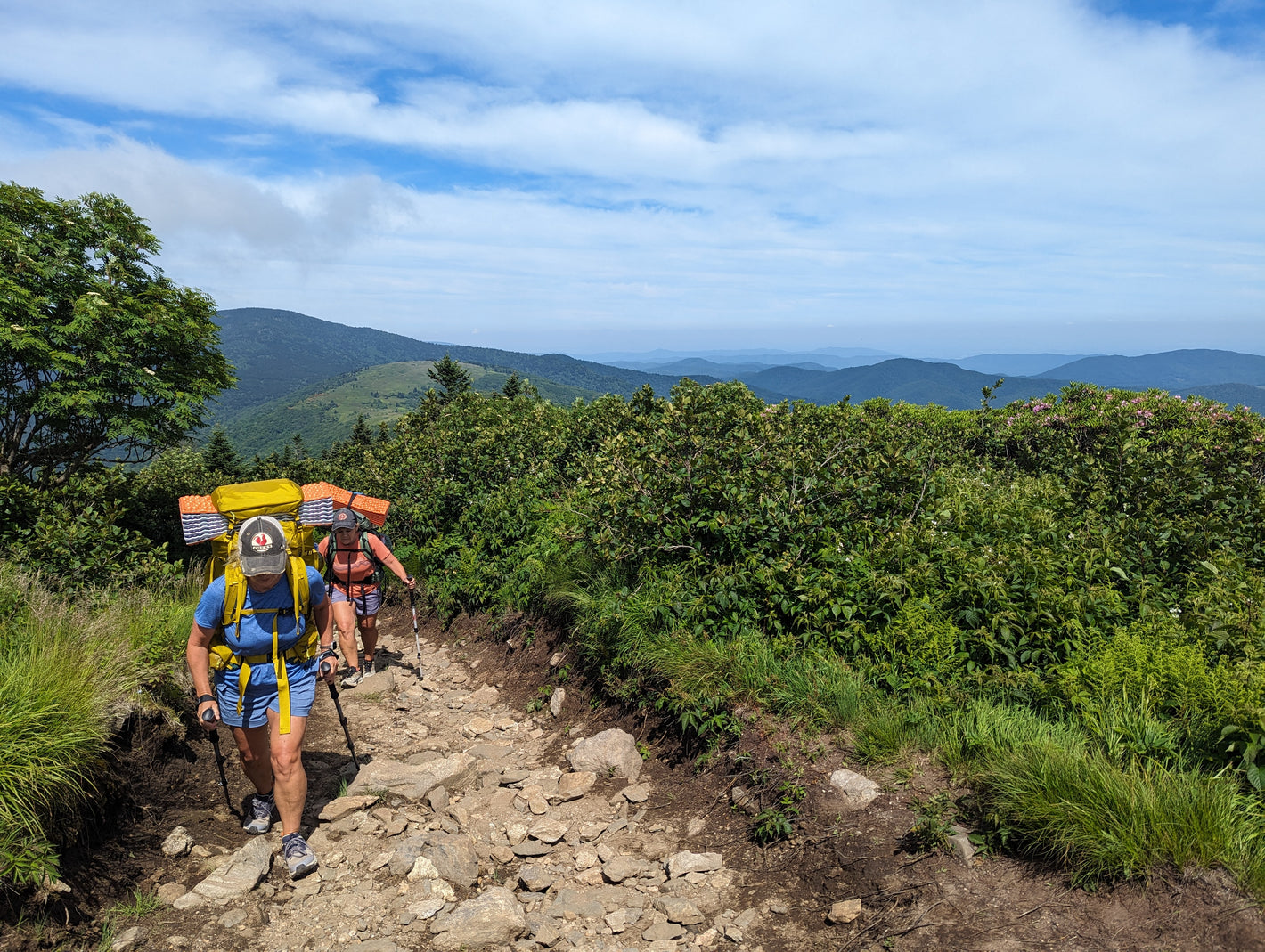 Two women with backpacks hiking on a rocky trail with mountains in the background