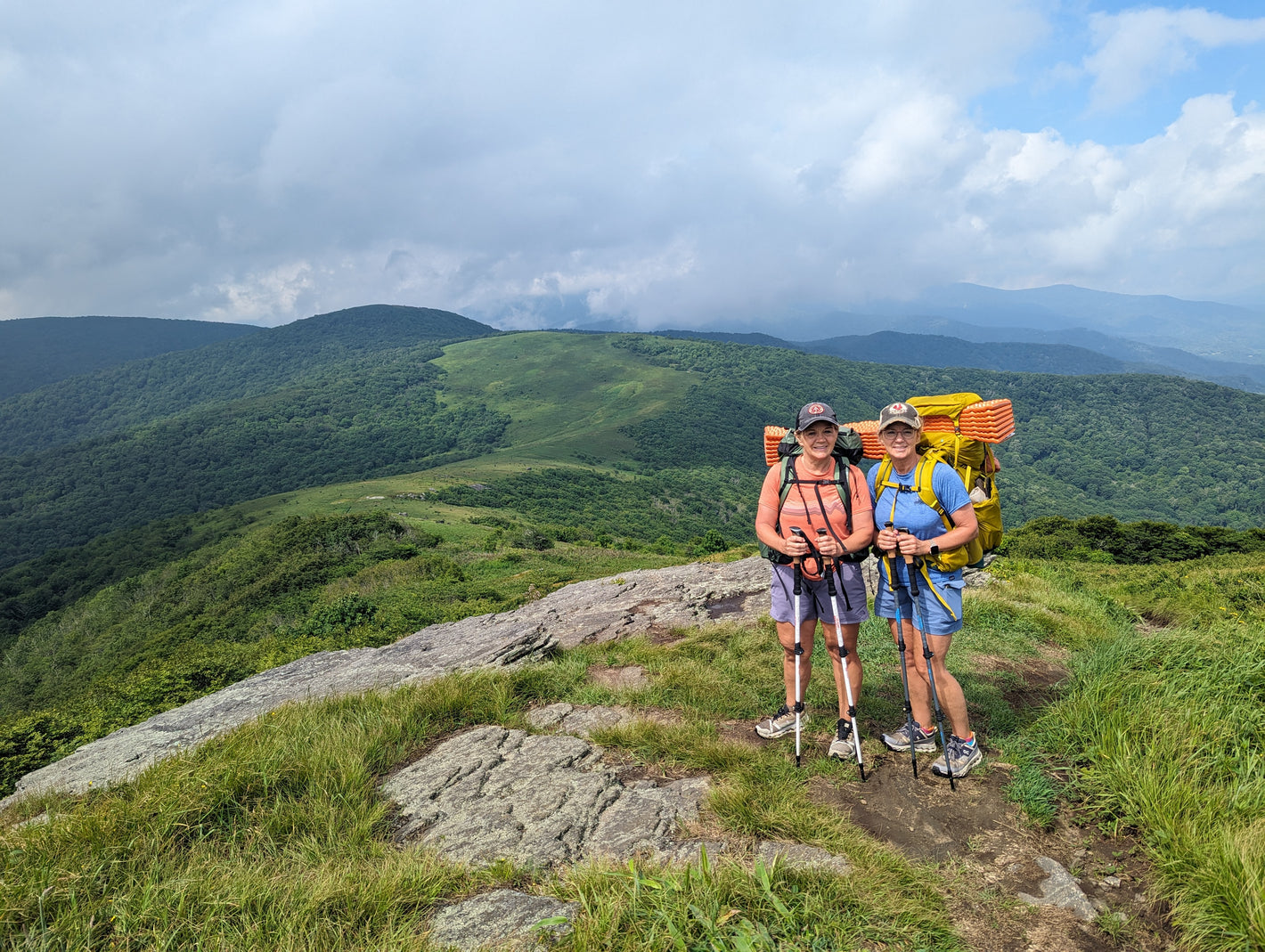 Two women with backpacks standing on top of a mountain with rolling green hills in the background