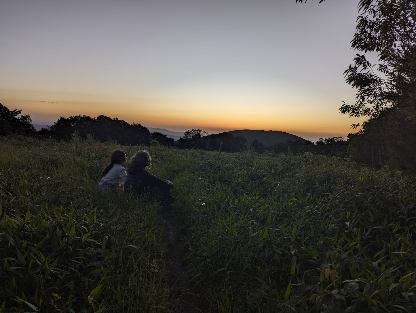 Two woman sitting in a grassy field watching the sunset over the mountains