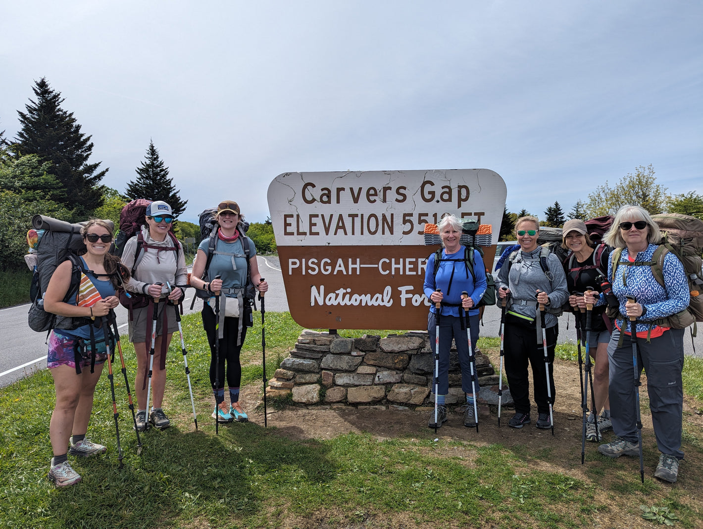 A group of seven women with backpacks and trekking poles standing in front of a sign for Carvers Gap