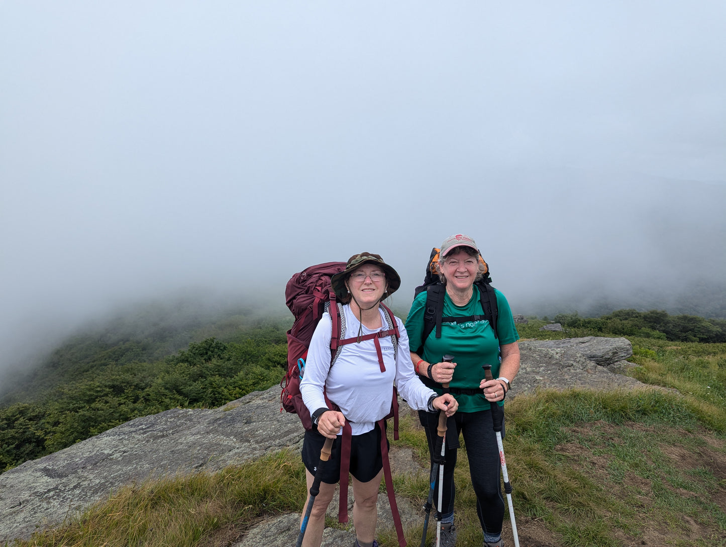 Two women with backpacks and trekking poles standing in a meadow with fog behind them