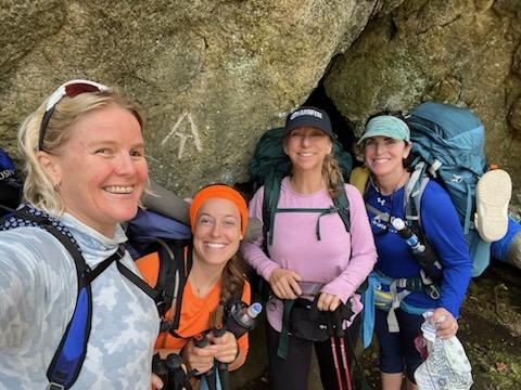 A group of women with backpacking gear in front of a rock with the Appalachian Trail symbol carved into it